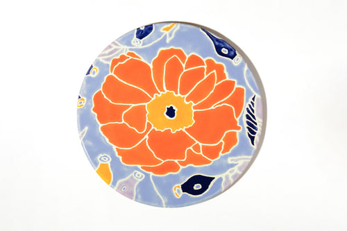 A round decorative plate or coaster featuring a stylized orange flower with a yellow center on a light blue background with small navy and yellow abstract accents.